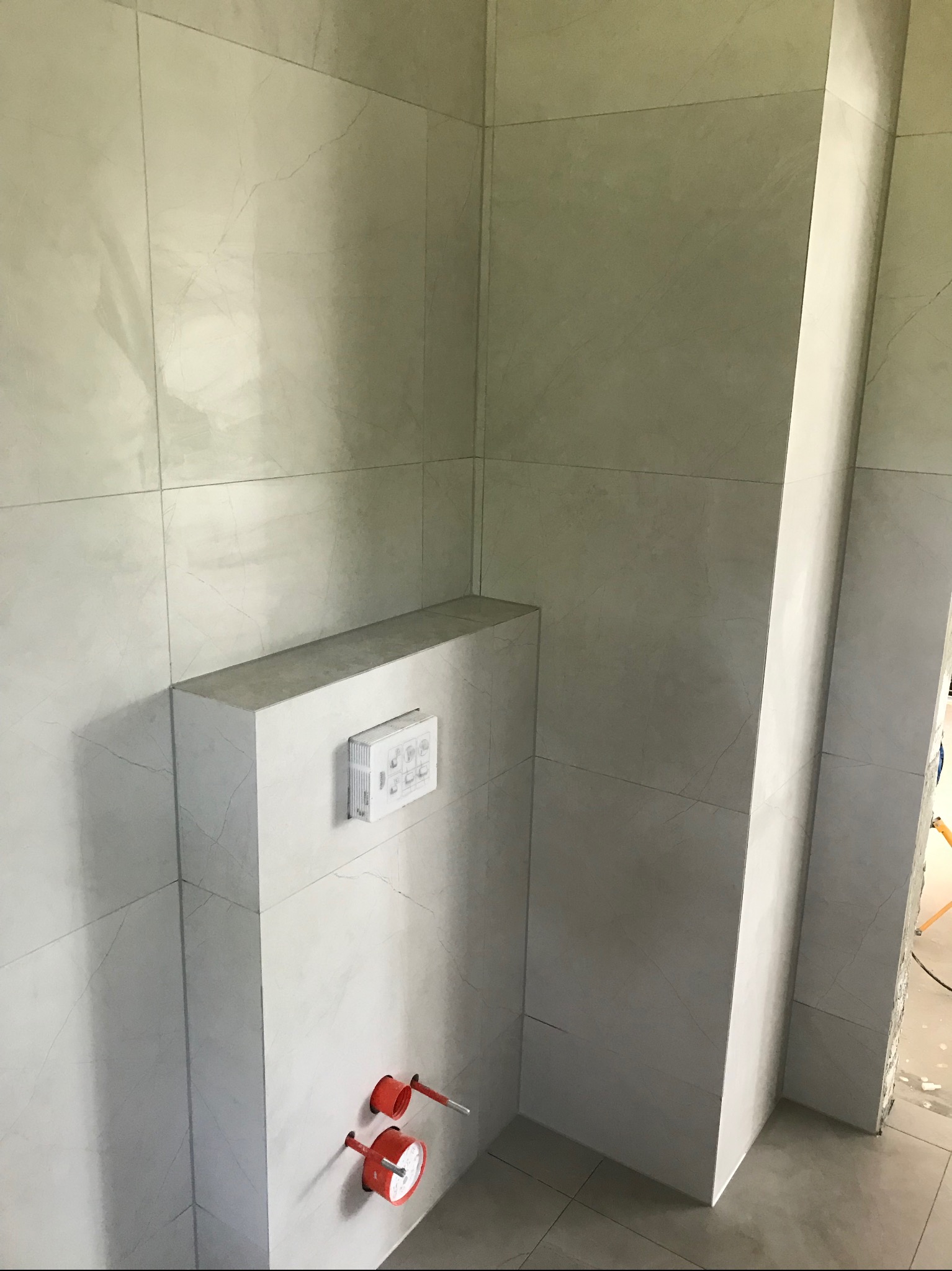 Wykończona łazienka z szarymi płytkami imitującymi marmur, widoczna zabudowa stelaża WC z przyciskiem spłukującym i wystającymi elementami instalacji wodnej.