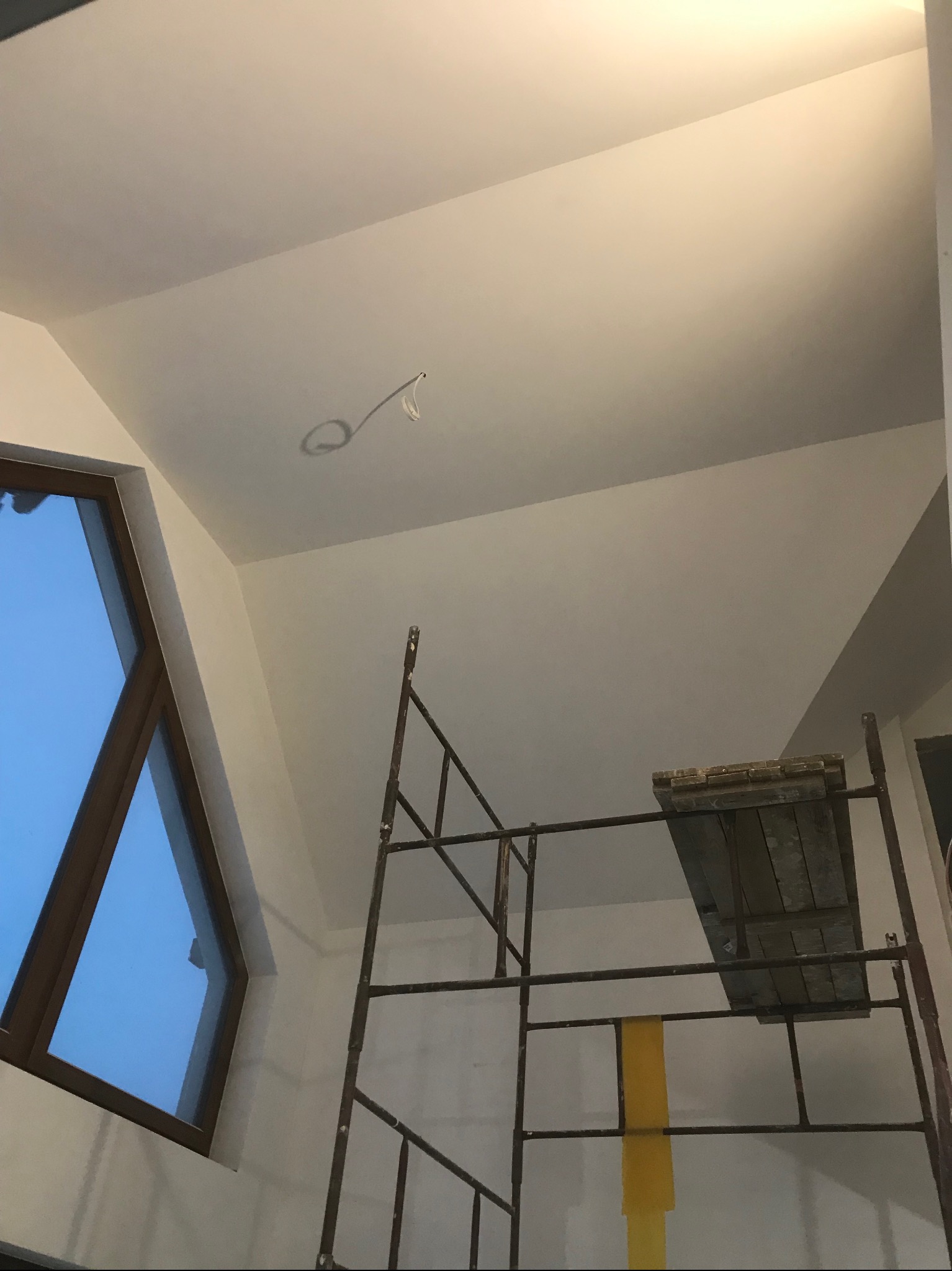 Białe wnętrze w trakcie prac wykończeniowych: widoczne rusztowanie, okno o nietypowym kształcie i gładka, pomalowana na biało powierzchnia sufitu z wystającym kablem.