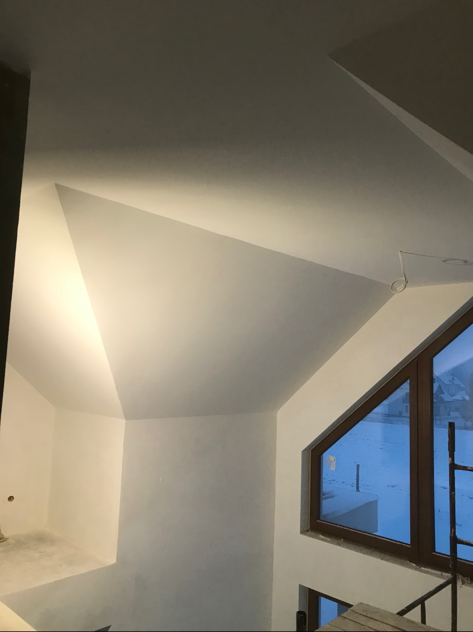 Wnętrze poddasza po gładziowaniu i malowaniu na biało, z oknem w kształcie trapezu i widocznym fragmentem drabiny. Ściany gładkie, gotowe do dalszych prac wykończeniowych.