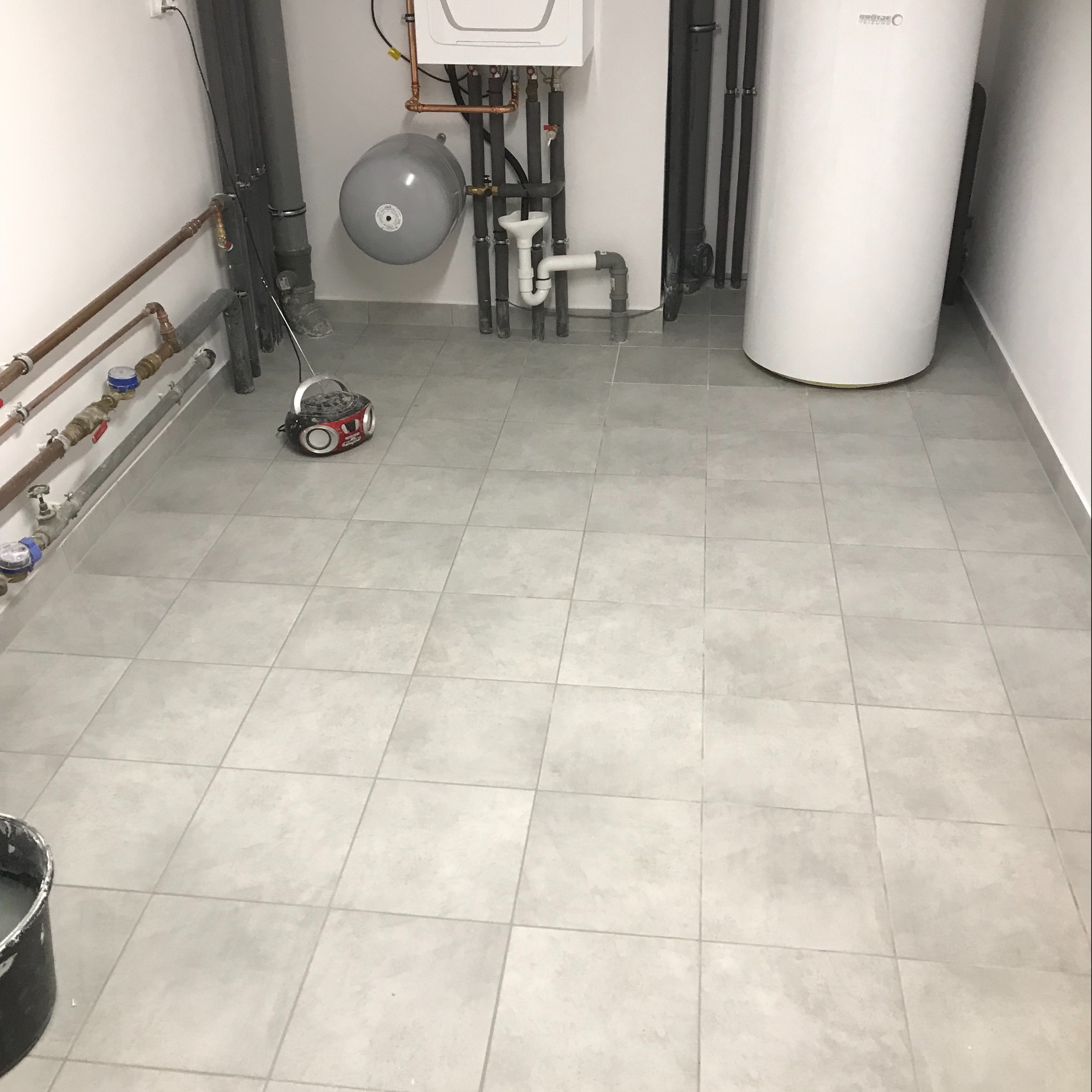 Wykończone pomieszczenie techniczne z szarymi płytkami podłogowymi, białym bojlerem, kotłem gazowym, instalacją hydrauliczną i grzewczą oraz radiem budowlanym na podłodze.