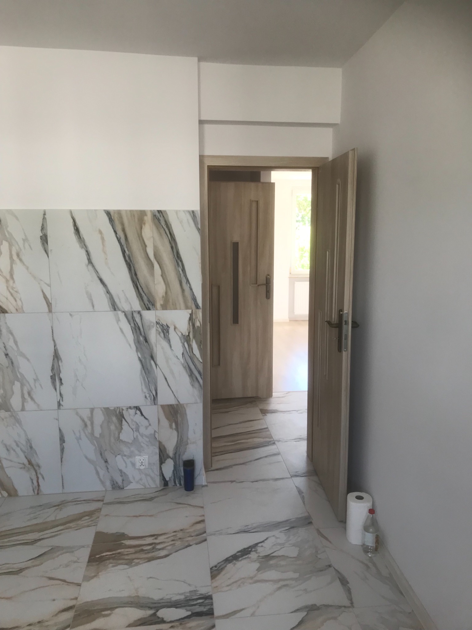 Wykończone wnętrze: białe ściany, płytki imitujące marmur na ścianie i podłodze, otwarte drzwi do kolejnego pomieszczenia, rolka papieru i butelka przy ścianie.