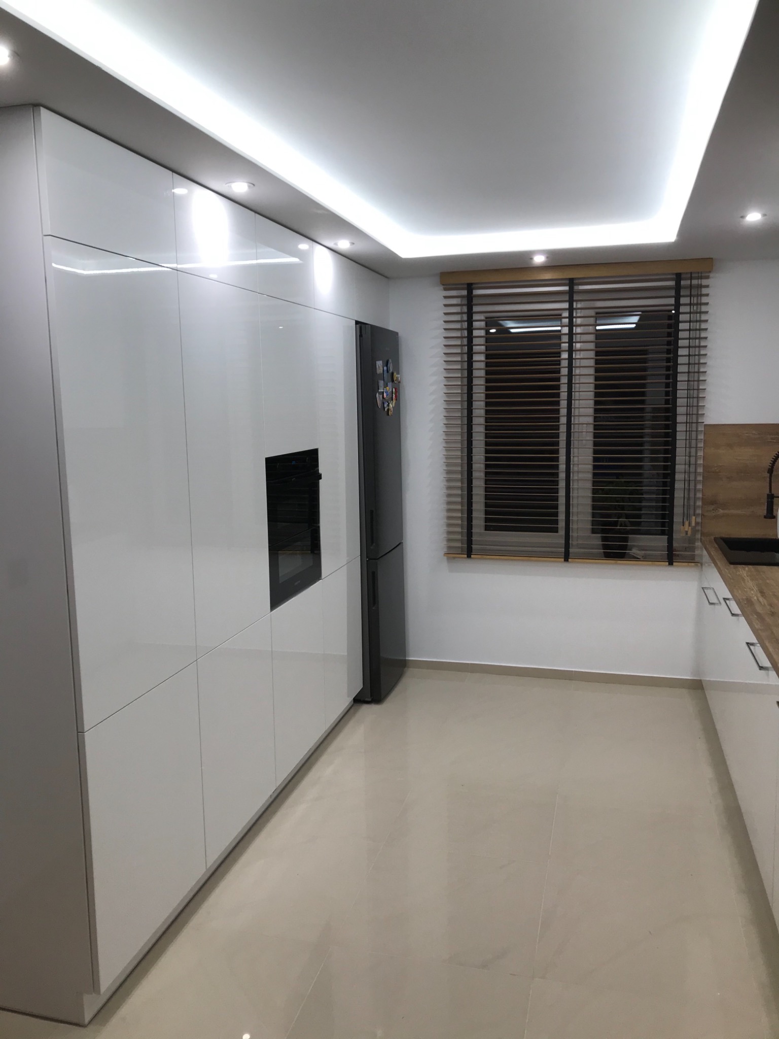 Nowoczesna kuchnia z białymi, lakierowanymi szafkami, wbudowanym piekarnikiem i czarną lodówką, oświetlona listwą LED pod sufitem, z beżową podłogą i oknem z drewnianymi żaluzjami.