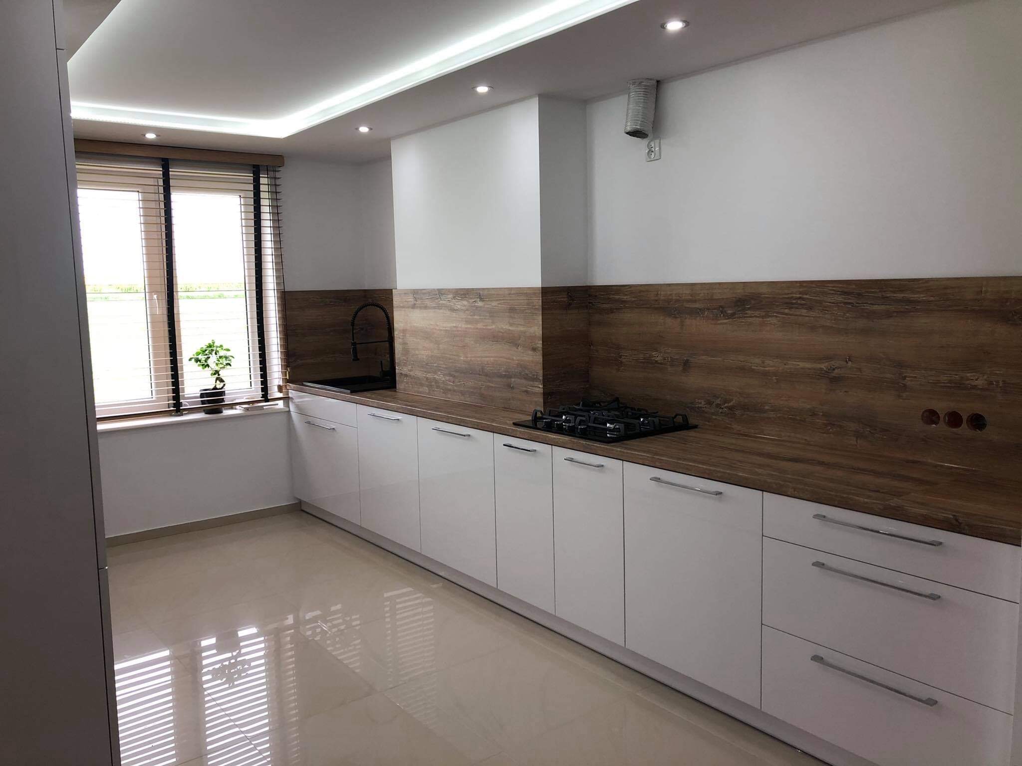 Nowoczesna, jasna kuchnia z białymi szafkami i drewnopodobnym blatem, oświetleniem LED podszafkowym i błyszczącą podłogą. Na blacie kuchenka gazowa. Widok na okno z żaluzjami i małą rośliną.