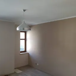 Pokój po malowaniu na beżowo-szaro, widoczne dwa okna z drewnianą ramą, podłoga w panelach oraz srebrna lampa w kształcie kropli zwisająca z białego sufitu.