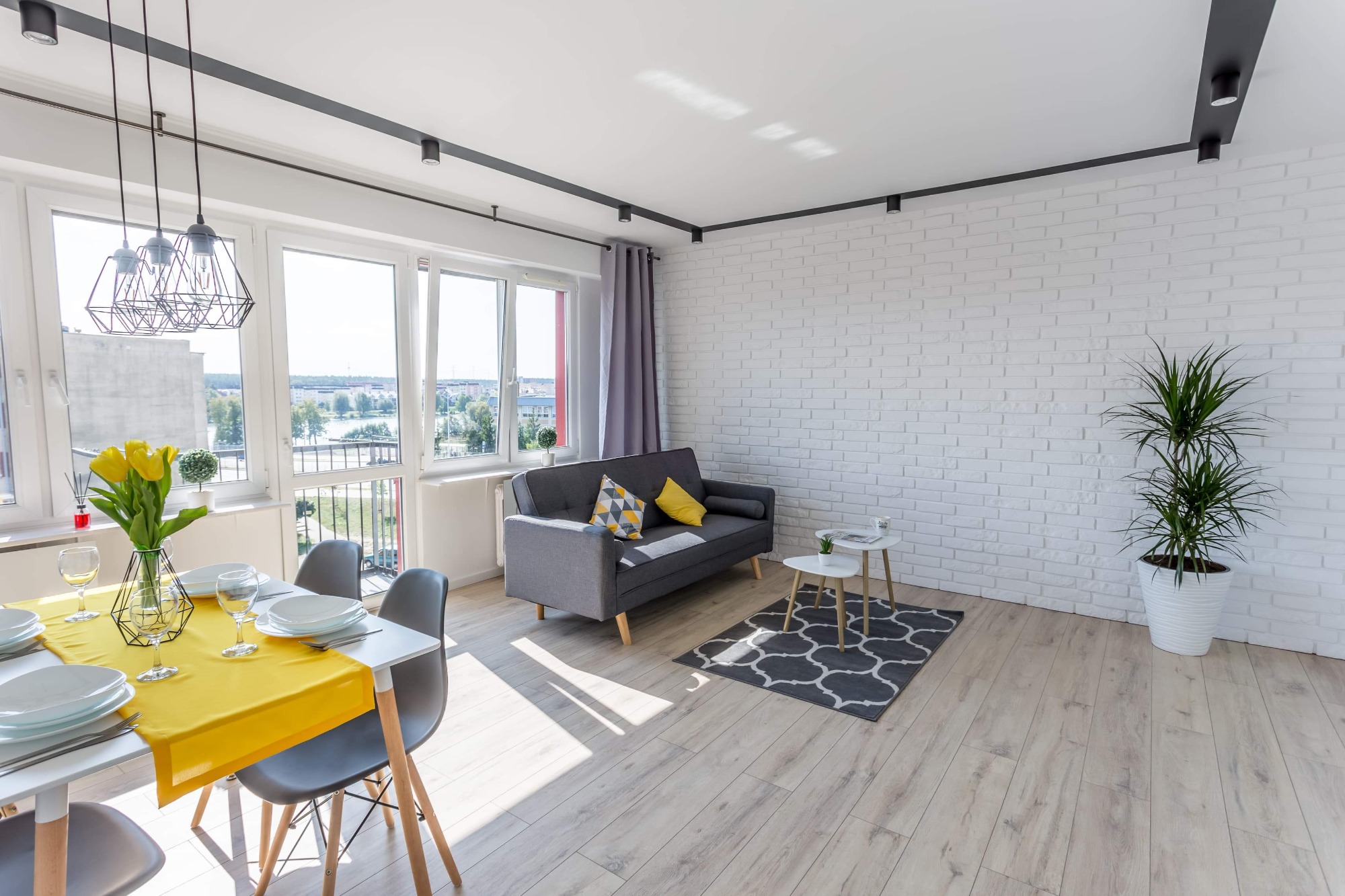 Jasne, nowocześnie urządzone wnętrze mieszkania z białymi cegłami na ścianie, szarą sofą z kolorowymi poduszkami, stołem z żółtym bieżnikiem i designerskimi lampami wiszącymi.