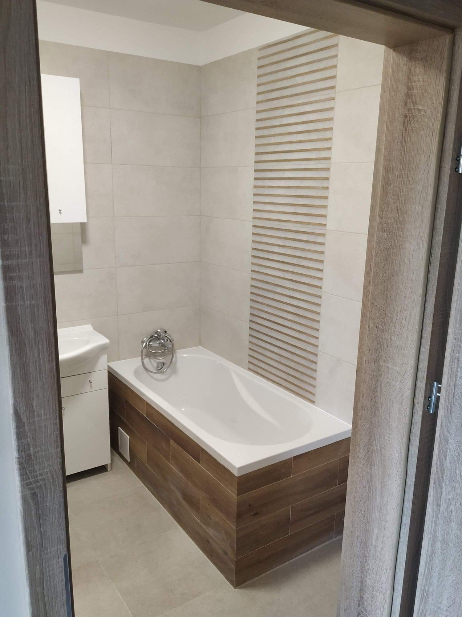 Nowoczesna łazienka z wanną obudowaną panelami imitującymi drewno, jasne płytki na ścianach z dekoracyjnym pasem poziomych pasków i biała szafka z umywalką, widok zza framugi drzwi.