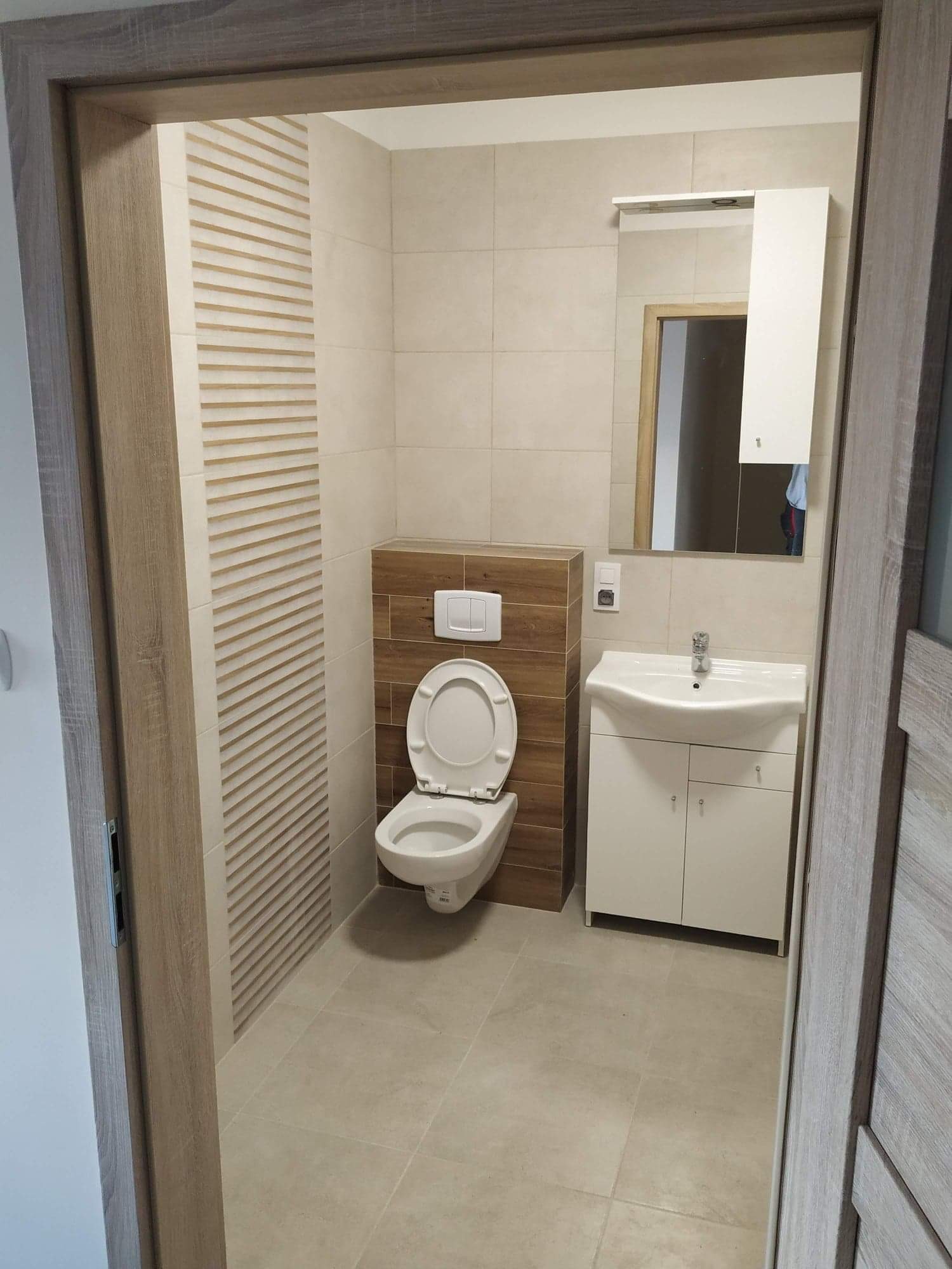 Jasna łazienka po renowacji, widok na toaletę z deską podniesioną, umywalkę z szafką, lustro z szafką wiszącą i dekoracyjne panele ścienne z poziomymi żłobieniami, całość utrzymana w beżowej...