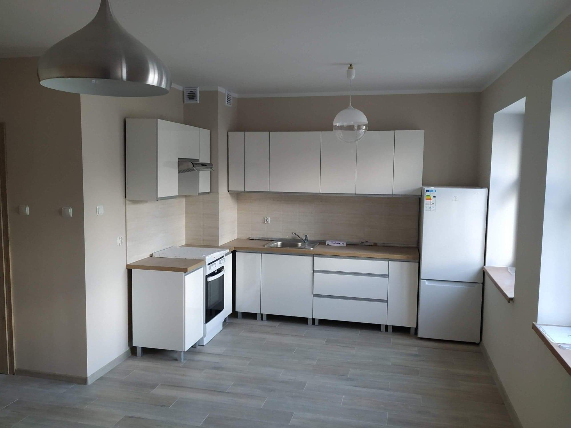 Jasna kuchnia po remoncie z białymi meblami, drewnianym blatem i szarą podłogą z płytek imitujących drewno, oświetlona dwoma lampami wiszącymi o różnych kształtach.