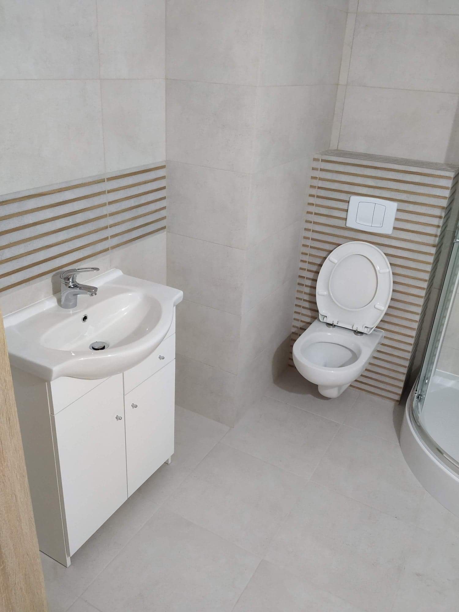 Jasna łazienka po remoncie: biała umywalka z szafką, toaleta z podwieszanym spłukiwaniem i fragment kabiny prysznicowej, płytki w odcieniach szarości z dekoracyjnym pasem z poziomych pasków...