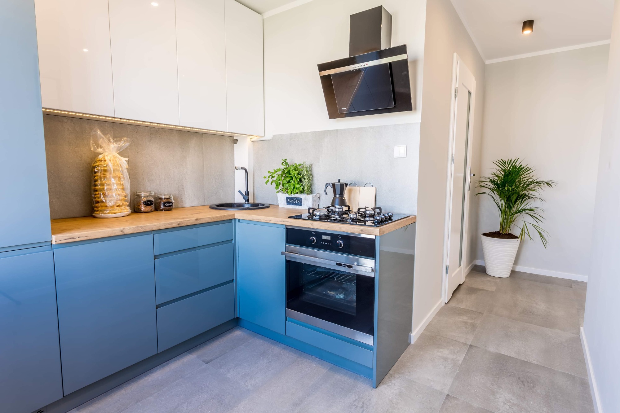 Nowoczesna, jasna kuchnia z błękitnymi szafkami, drewnianym blatem, czarnym zlewem i płytą grzewczą. Na blacie zioła w doniczce i kawiarka. Na ścianie szara płytka imitująca beton. W tle widoczny...