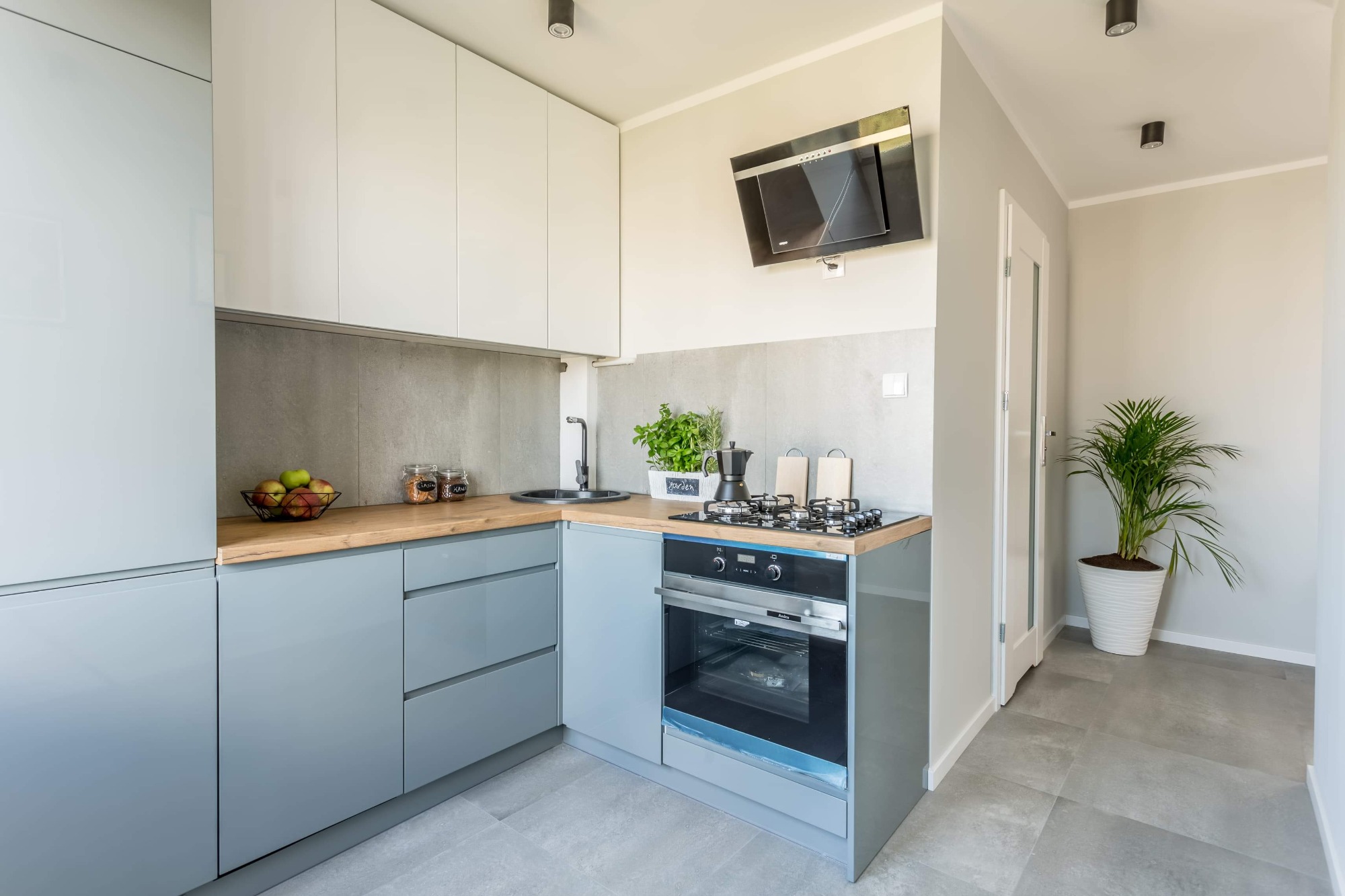 Jasna kuchnia w odcieniach szarości i bieli z drewnianym blatem, czarną armaturą, płytą gazową i piekarnikiem marki Amica. Na blacie zioła w doniczce i włoska kafetierka.