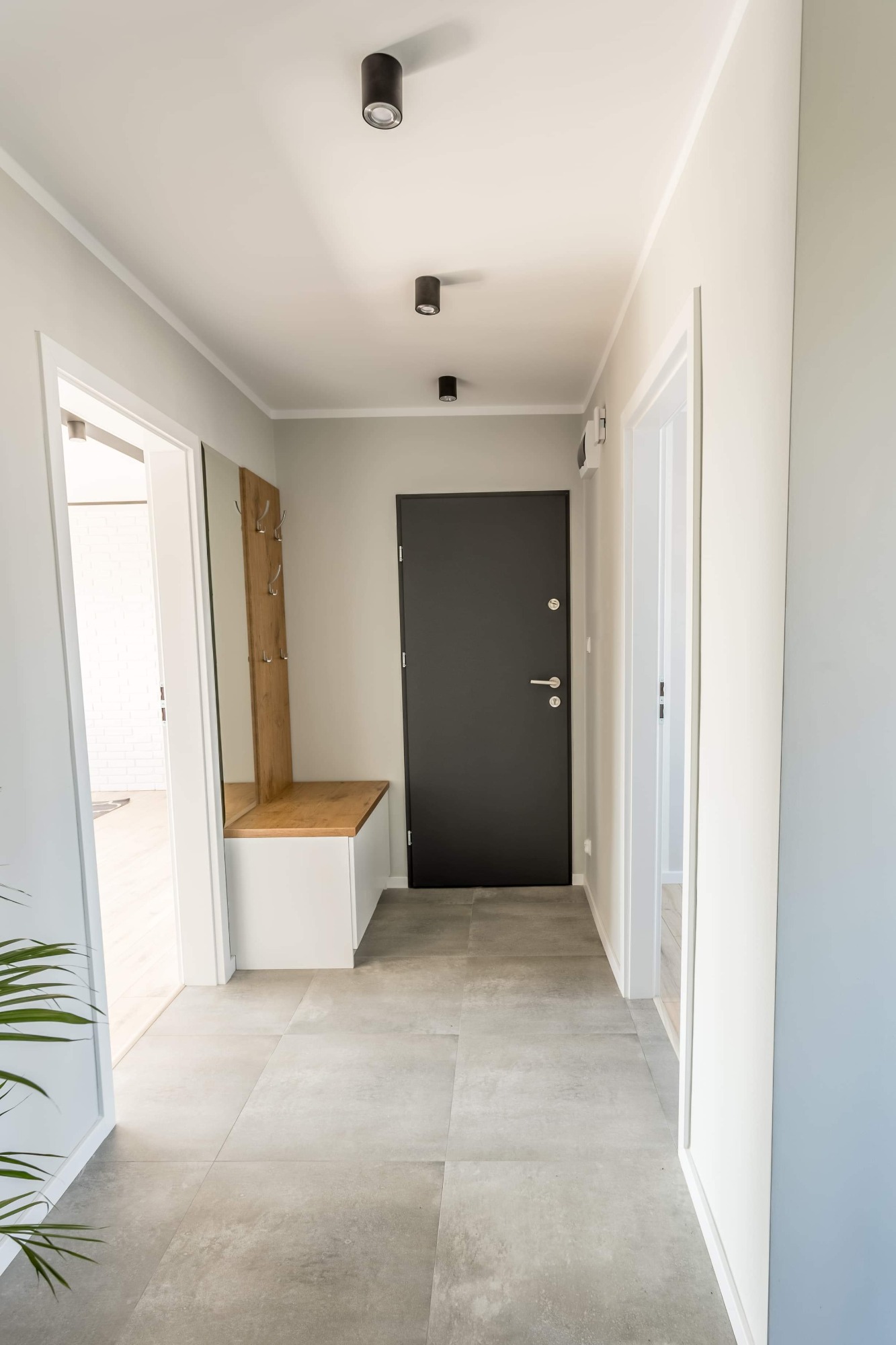 Jasny korytarz z szarą podłogą z płytek, czarnymi lampami sufitowymi, ciemnymi drzwiami wejściowymi i szafką z wieszakami na ścianie.