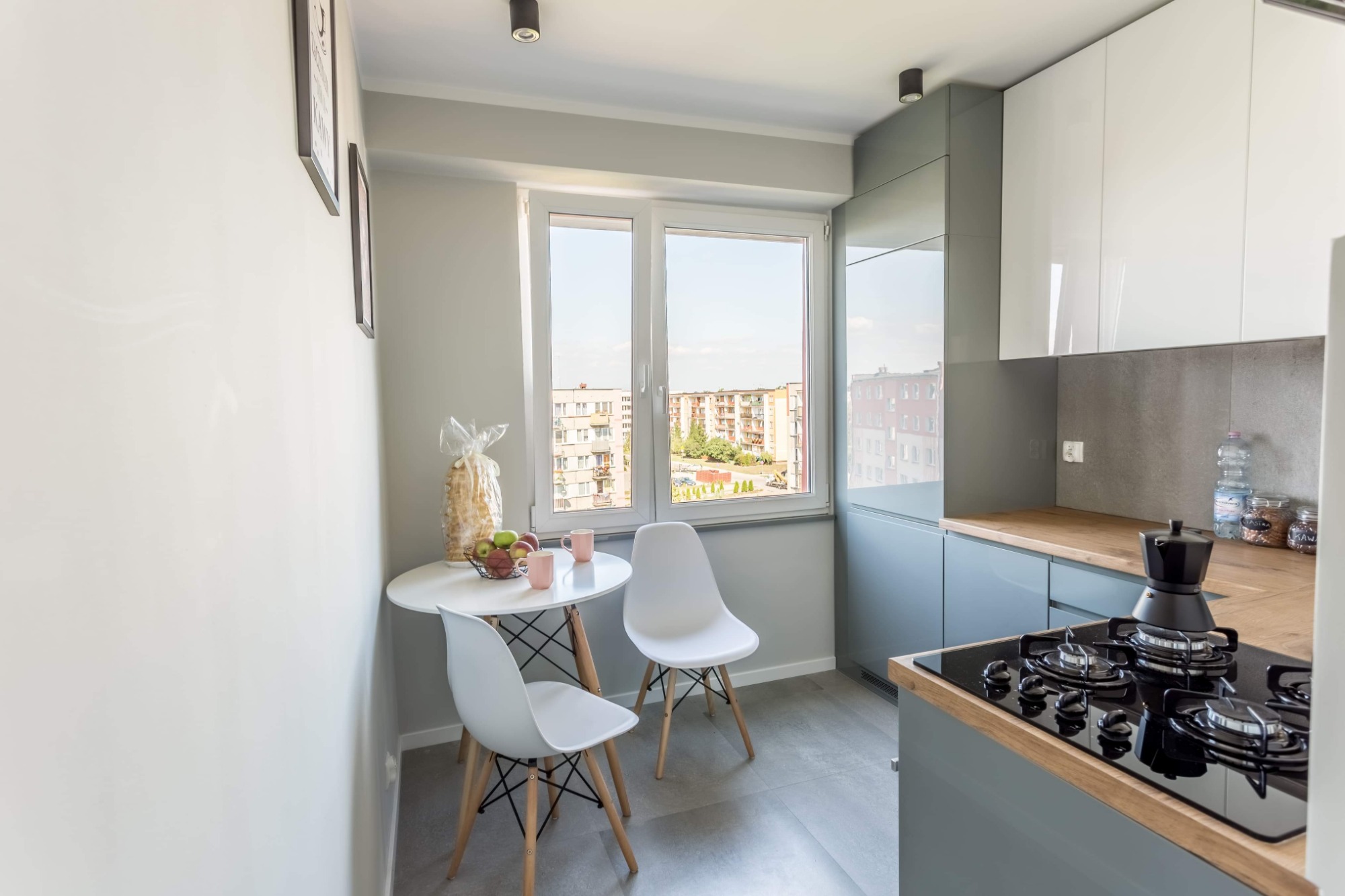 Nowoczesna kuchnia w szaro-białej kolorystyce z drewnianym blatem, okrągłym stołem z owocami i widokiem na osiedle z okna.