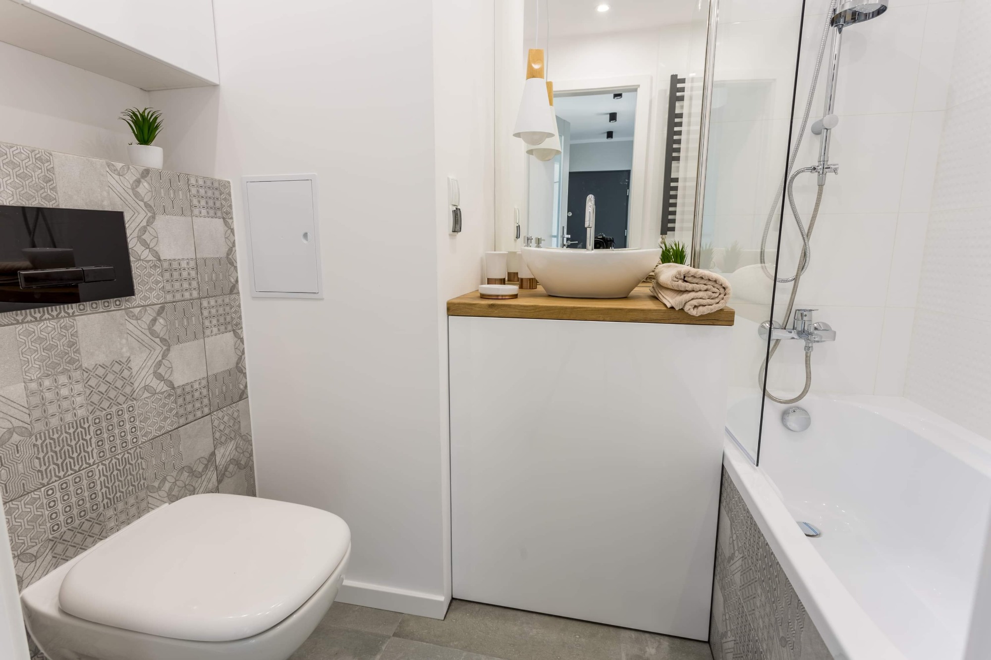 Nowoczesna łazienka z wiszącą toaletą, umywalką nablatową na drewnianym blacie, kabiną prysznicową i dekoracyjnymi płytkami w odcieniach szarości.