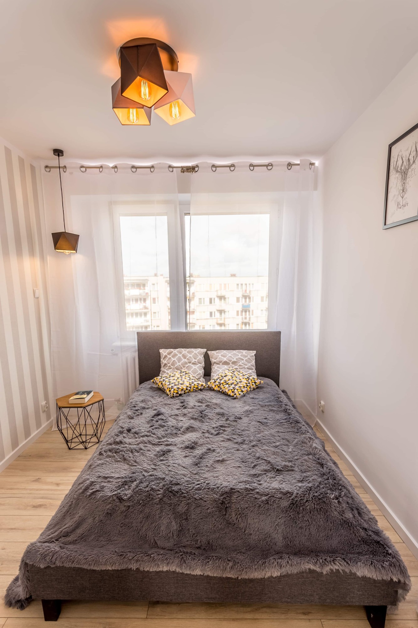 Jasna sypialnia z szarym łóżkiem przykrytym puszystą narzutą, geometryczne poduszki, lampa wisząca z trzema kloszami w kształcie rombów, mały stolik nocny z książką, okno z białymi zasłonami.