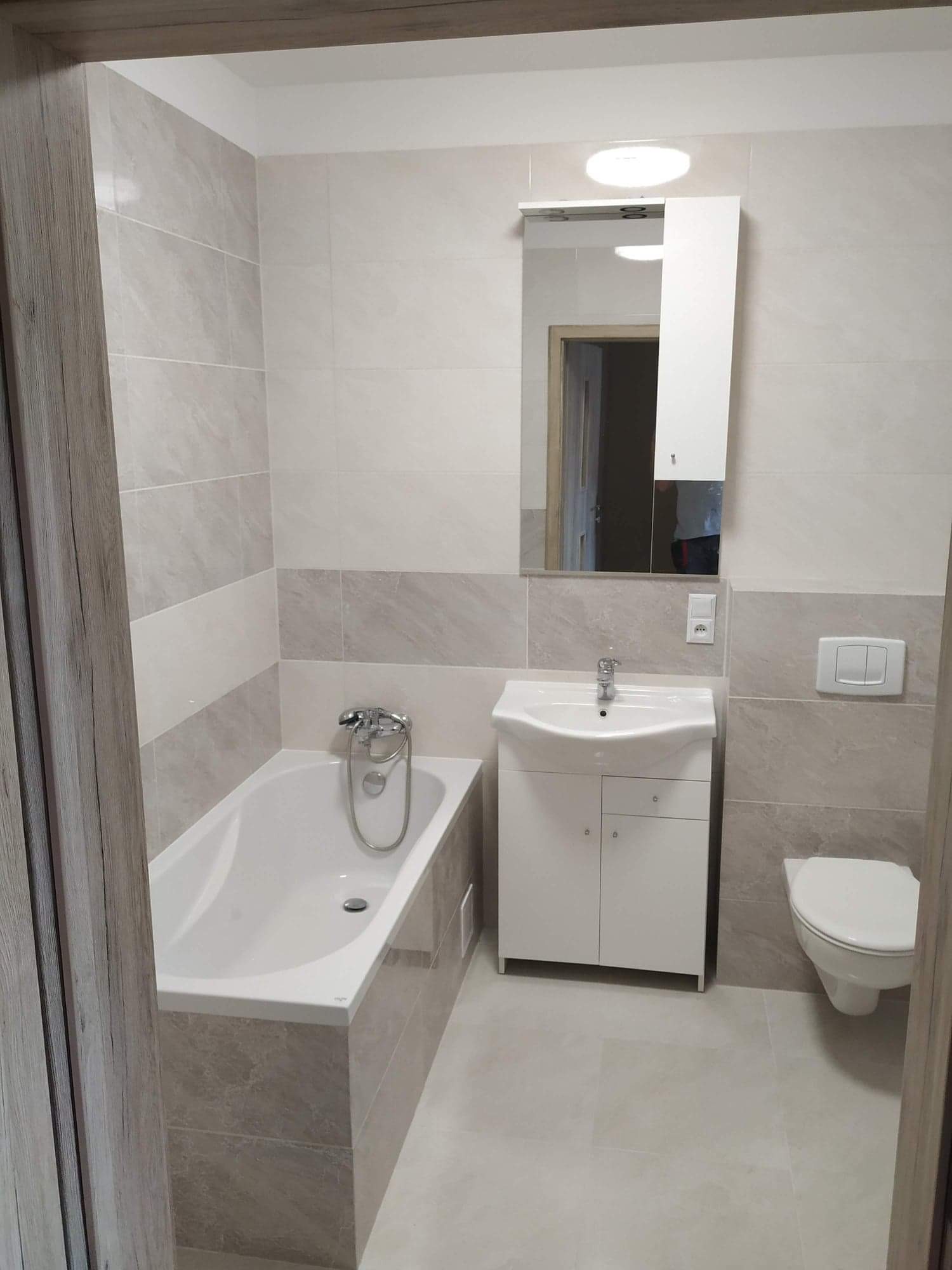 Jasna łazienka po remoncie: beżowe płytki imitujące marmur na ścianach i podłodze, biała wanna z chromowaną baterią, umywalka z szafką i lustro z oświetleniem, wisząca toaleta z podwójnym...