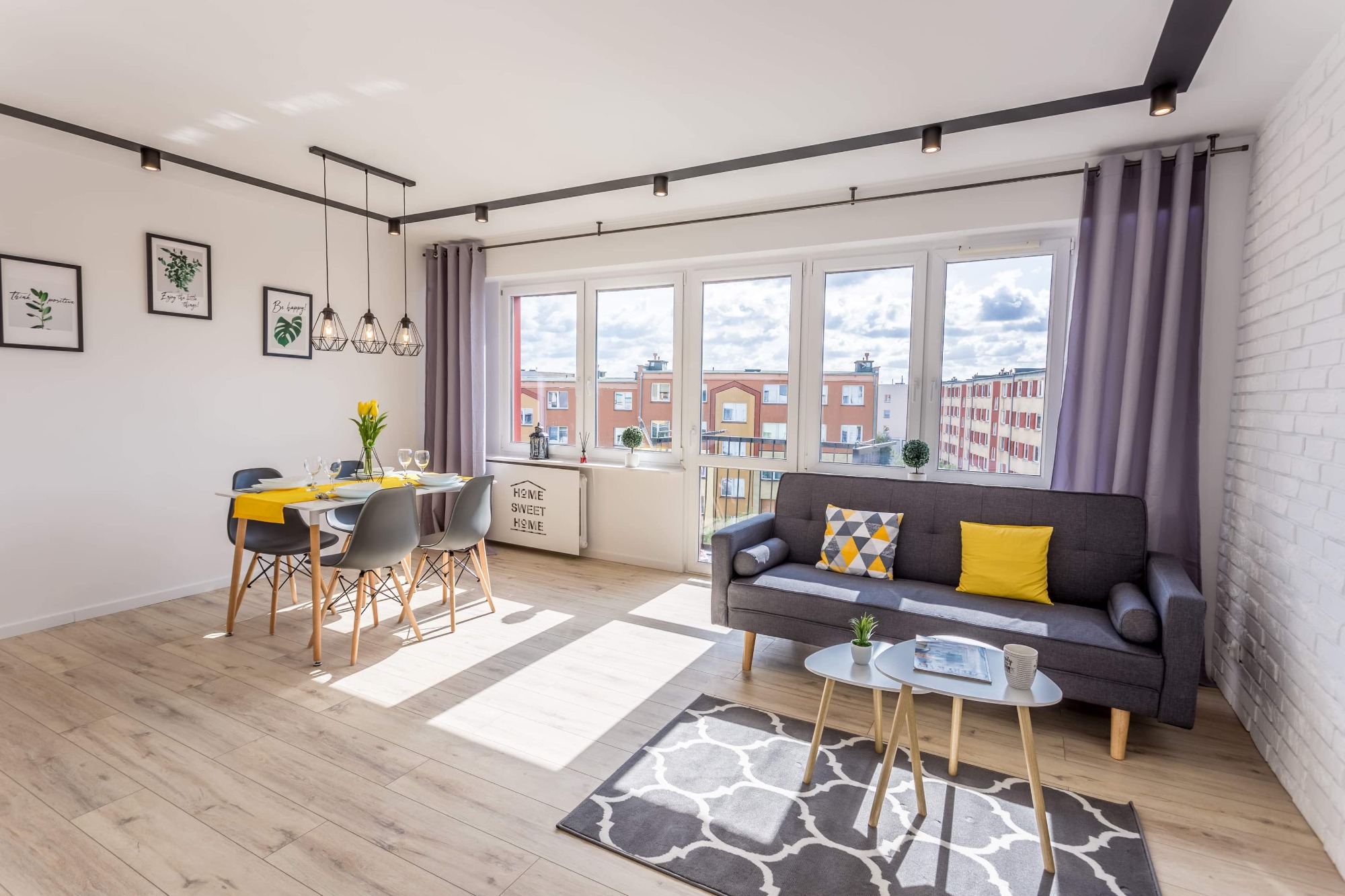 Jasne wnętrze mieszkania z drewnianą podłogą, szarą sofą z żółtymi poduszkami, białym stołem z szarymi krzesłami i żółtym obrusem oraz oknami z widokiem na miasto. Na ścianie biała cegła i czarne...