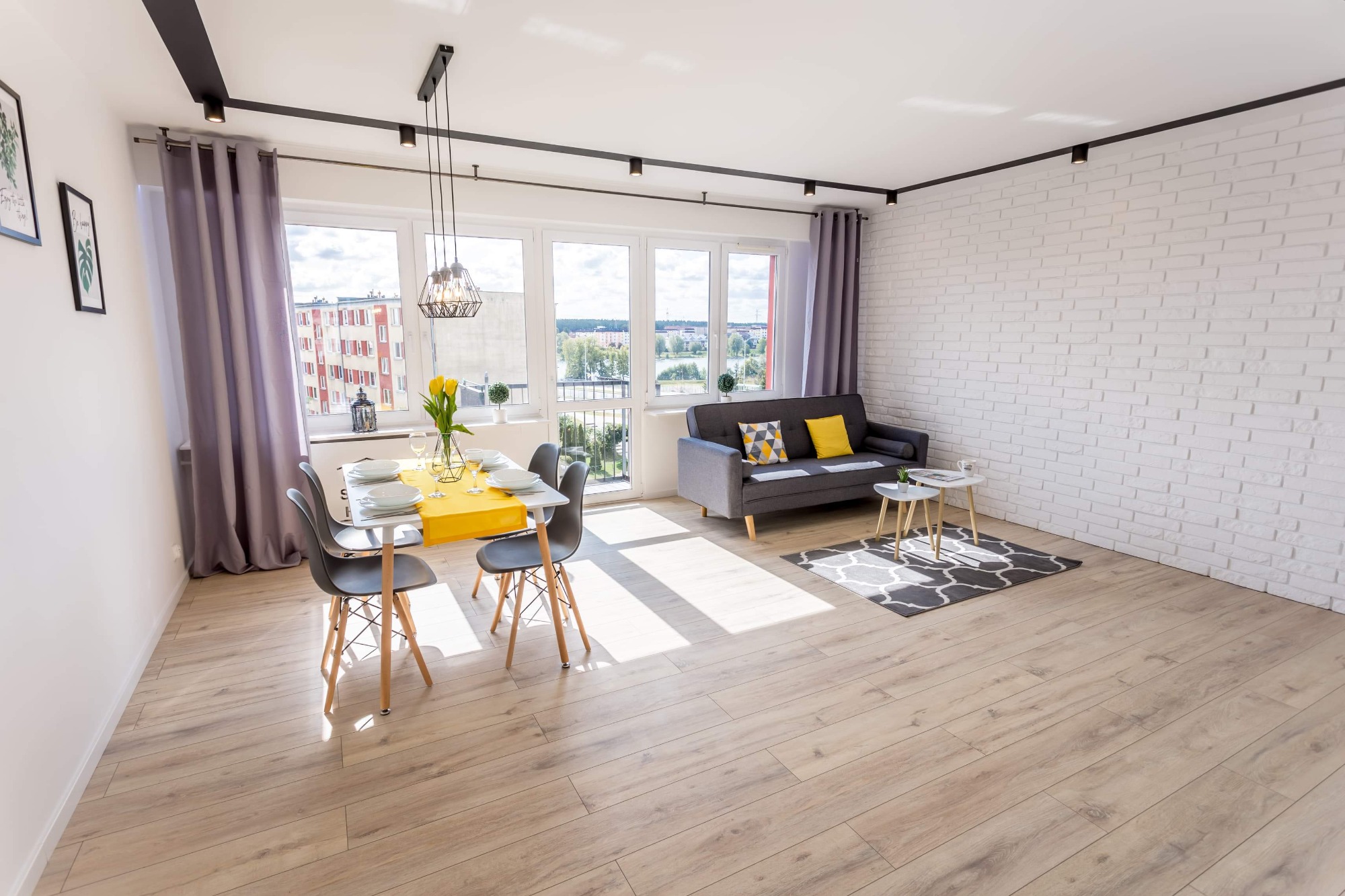 Jasne wnętrze mieszkania z drewnianą podłogą, białymi ścianami z cegły, szarą sofą z żółtymi poduszkami, stołem z żółtym bieżnikiem i bukietem tulipanów w wazonie, oświetlone punktowymi lampami...