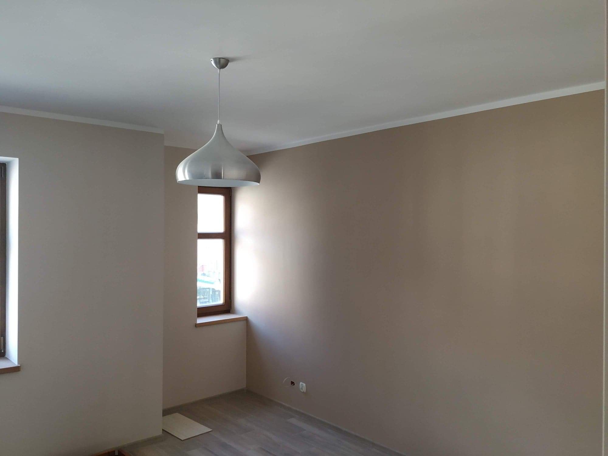 Pokój po malowaniu na beżowo-szaro, widoczne dwa okna z drewnianą ramą, podłoga w panelach oraz srebrna lampa w kształcie kropli zwisająca z białego sufitu.