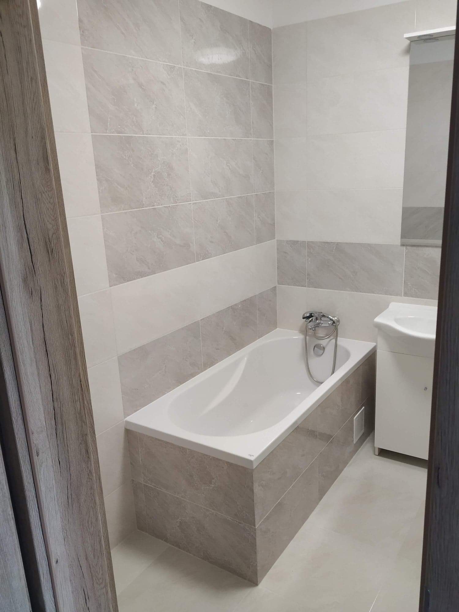 Wykończona łazienka w odcieniach szarości i bieli, z wanną obudowaną płytkami, umywalką z szafką i lustrem.