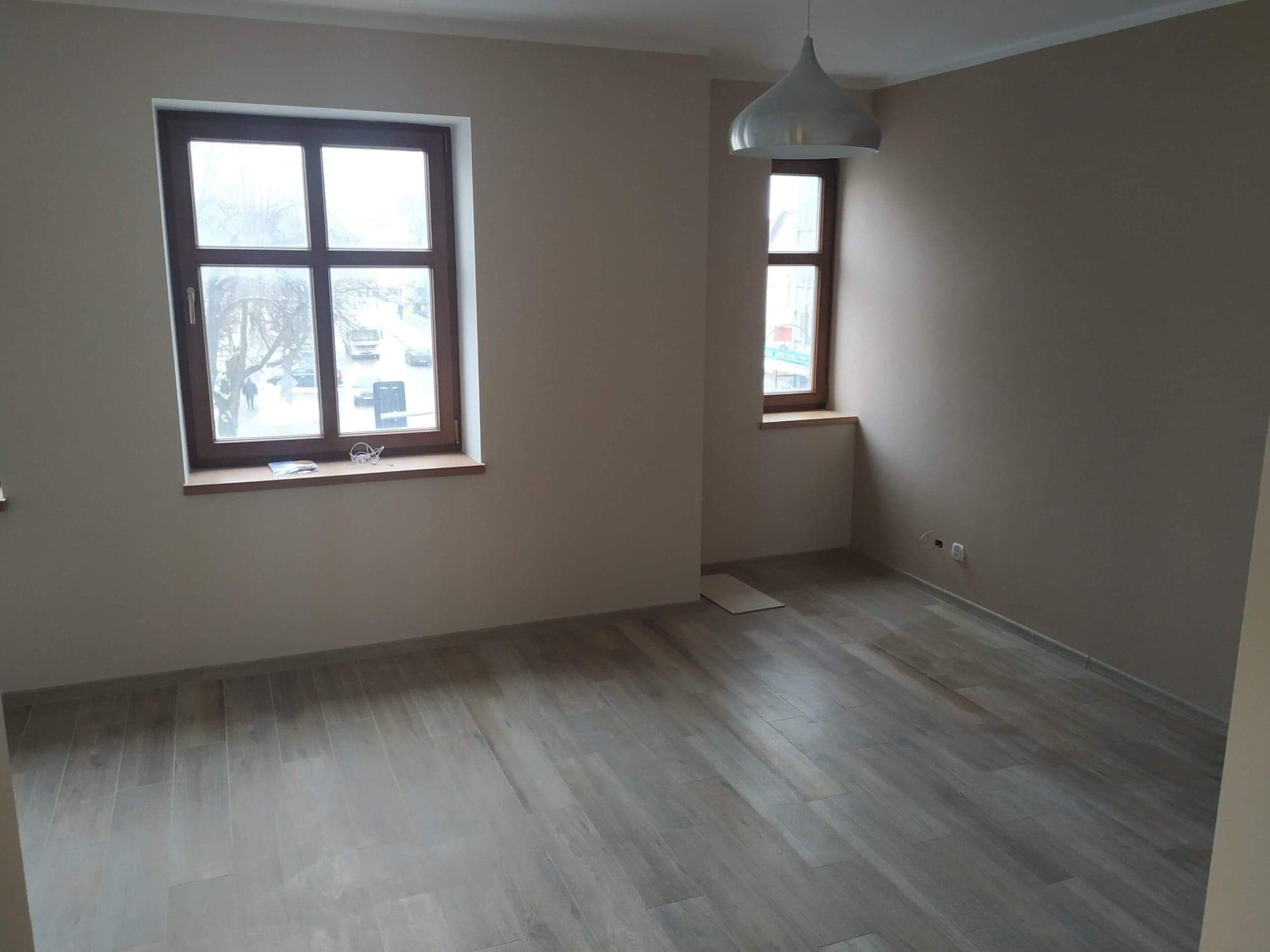 Puste, jasne pomieszczenie po remoncie z drewnopodobną podłogą, dwoma oknami o różnej wielkości i nowoczesną lampą wiszącą.
