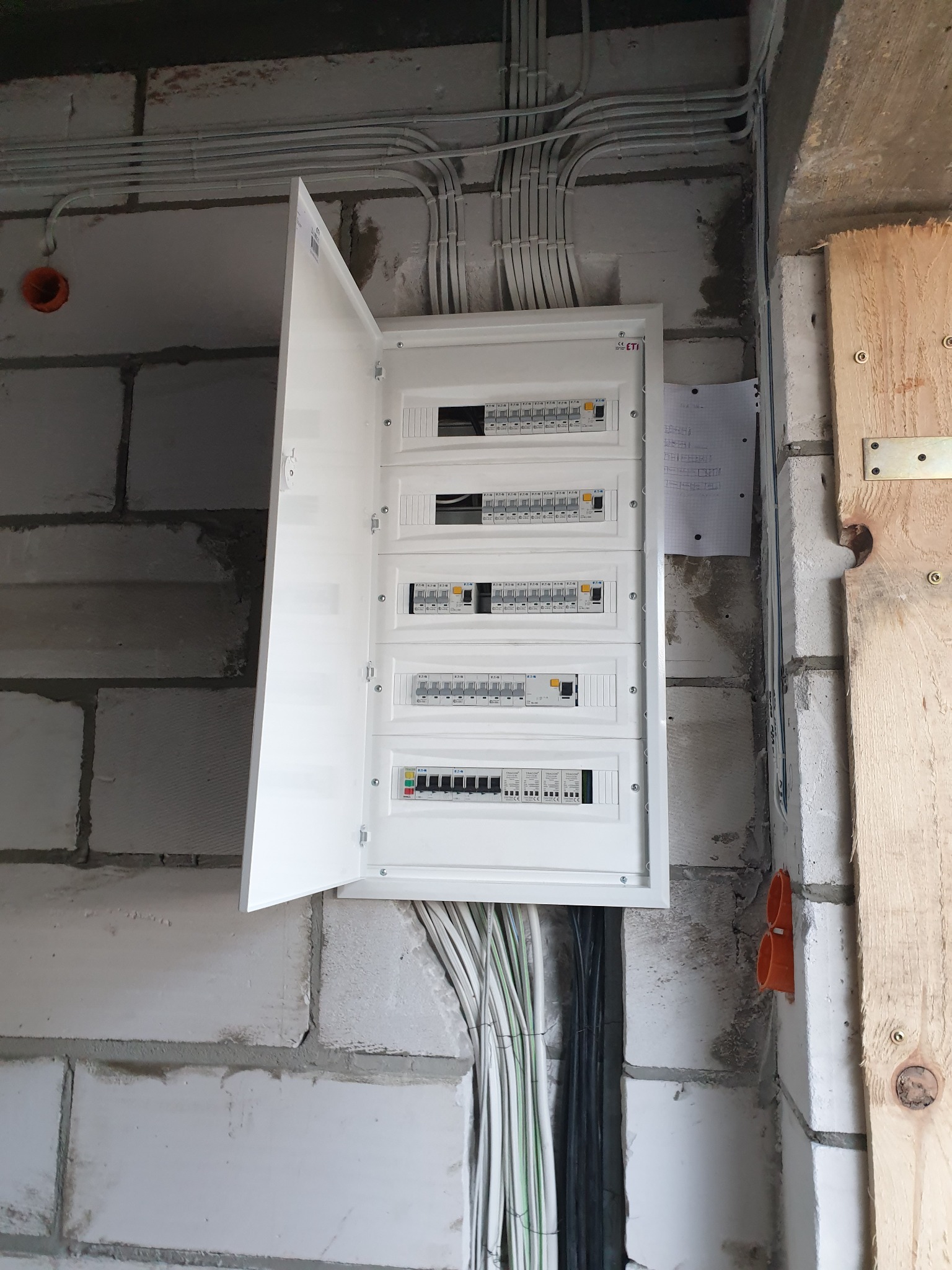 Otwarta skrzynka elektryczna z bezpiecznikami zamontowana na surowej ścianie z bloczków, widoczne kable w peszlach i puszki instalacyjne, kartka z notatkami obok skrzynki.