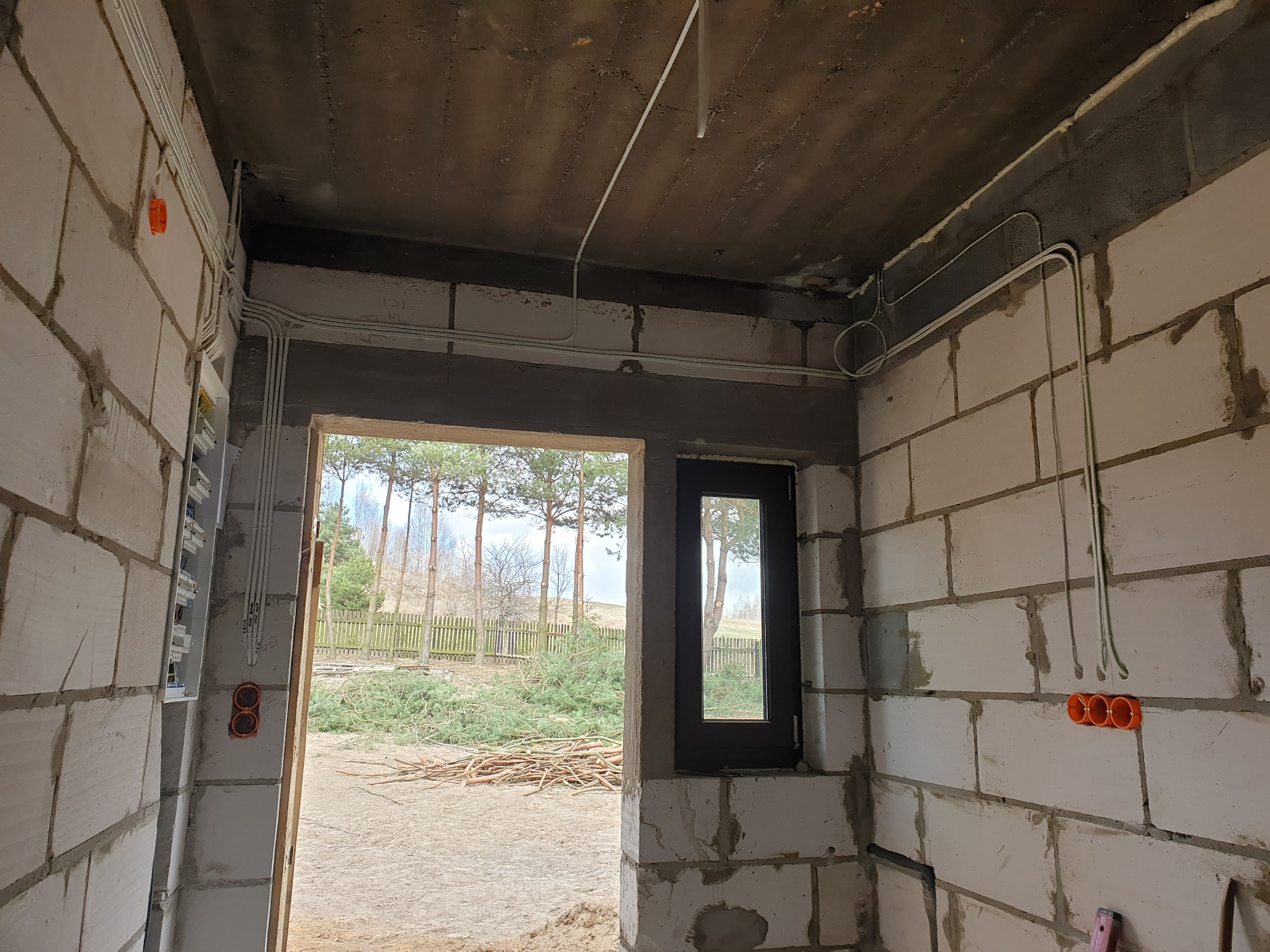 Surowy stan wnętrza budynku w trakcie prac, widoczne rozprowadzone kable elektryczne w peszlach na ścianach z betonu komórkowego, puszki instalacyjne, skrzynka bezpiecznikowa i przewody pod sufitem...