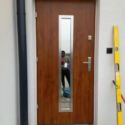 Drewniane drzwi wejściowe z przeszkleniem w metalowej ramie, widoczne narzędzia i odbicie osoby w szybie, poziomica oparta o ścianę.