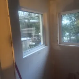 Dwa białe okna PCV zamontowane w surowym wnętrzu z otynkowanymi ścianami, widoczne ślady prac wykończeniowych.