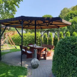 Altana ogrodowa z ciemnobrązowym dachem krytym gontem bitumicznym, otoczona zielenią i brukowanym placem, z drewnianym stołem i ławkami wewnątrz.
