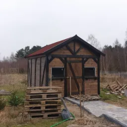 Drewniany domek narzędziowy z czerwonym dachem, czarnymi belkami i paletami przed wejściem, na tle lasu