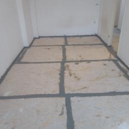 J.P. Kompleksowe Usługi Remontowe - Przygotowanie podłoża pod panele: płyty OSB z wypełnionymi szparami szarą masą uszczelniającą, widok z góry, surowe ściany w tle.