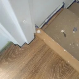 Fragment układania paneli podłogowych przy futrynie drzwiowej, widoczne niedoskonałości montażu i zabrudzenia farbą na ścianie.