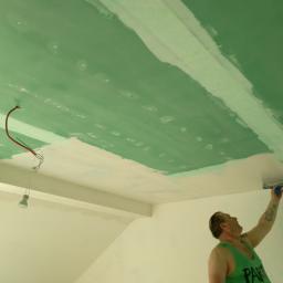 J.P. Kompleksowe Usługi Remontowe - Pracownik w zielonej koszulce szpachluje sufit w Gdańsku, widoczne niedokończone połączenia płyt gipsowo-kartonowych i wiszące przewody elektryczne z żarówką.