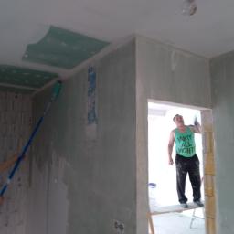 J.P. Kompleksowe Usługi Remontowe - Prace wykończeniowe w pomieszczeniu: mężczyzna w zielonym podkoszulku z napisem 'PARTY ALL NIGHT' stoi na rusztowaniu i wygładza ścianę, drugi mężczyzna maluje sufit wałkiem na długim kiju...