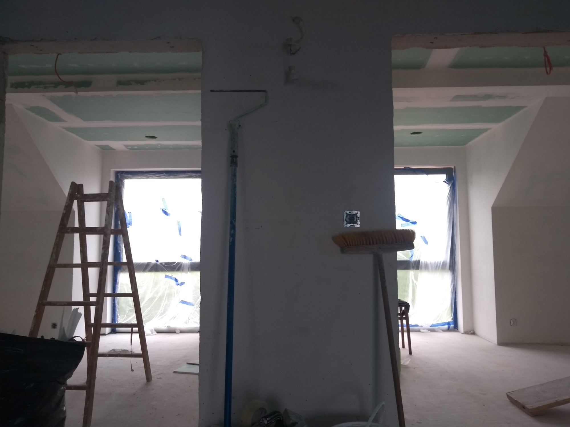 Wnętrze w trakcie remontu, widoczne szpachlowane ściany, sufit z płyt gipsowo-kartonowych, zabezpieczone okna folią, drabina, wałek malarski i szczotka oparta o ścianę.