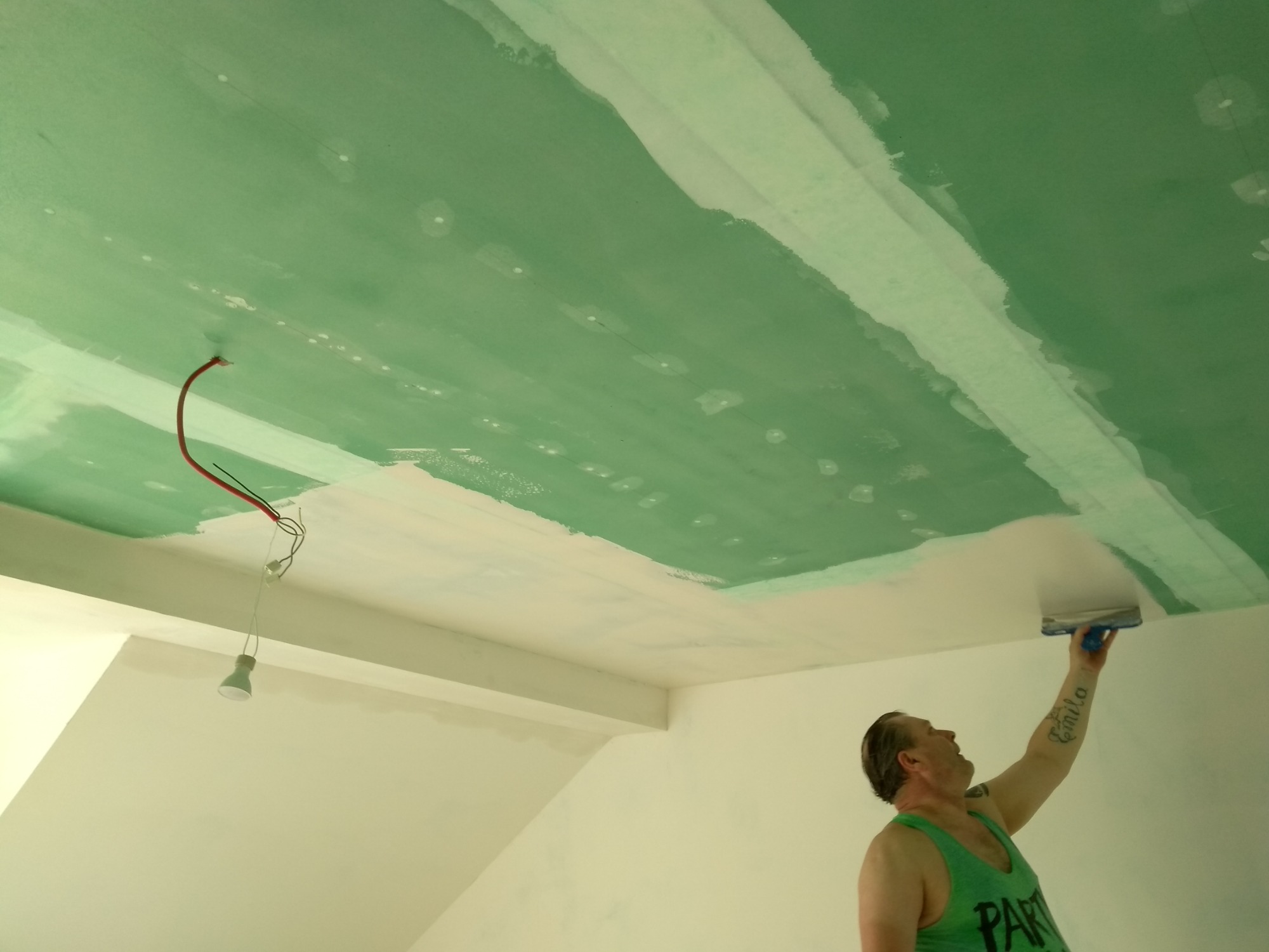 Pracownik w zielonej koszulce szpachluje sufit w Gdańsku, widoczne niedokończone połączenia płyt gipsowo-kartonowych i wiszące przewody elektryczne z żarówką.