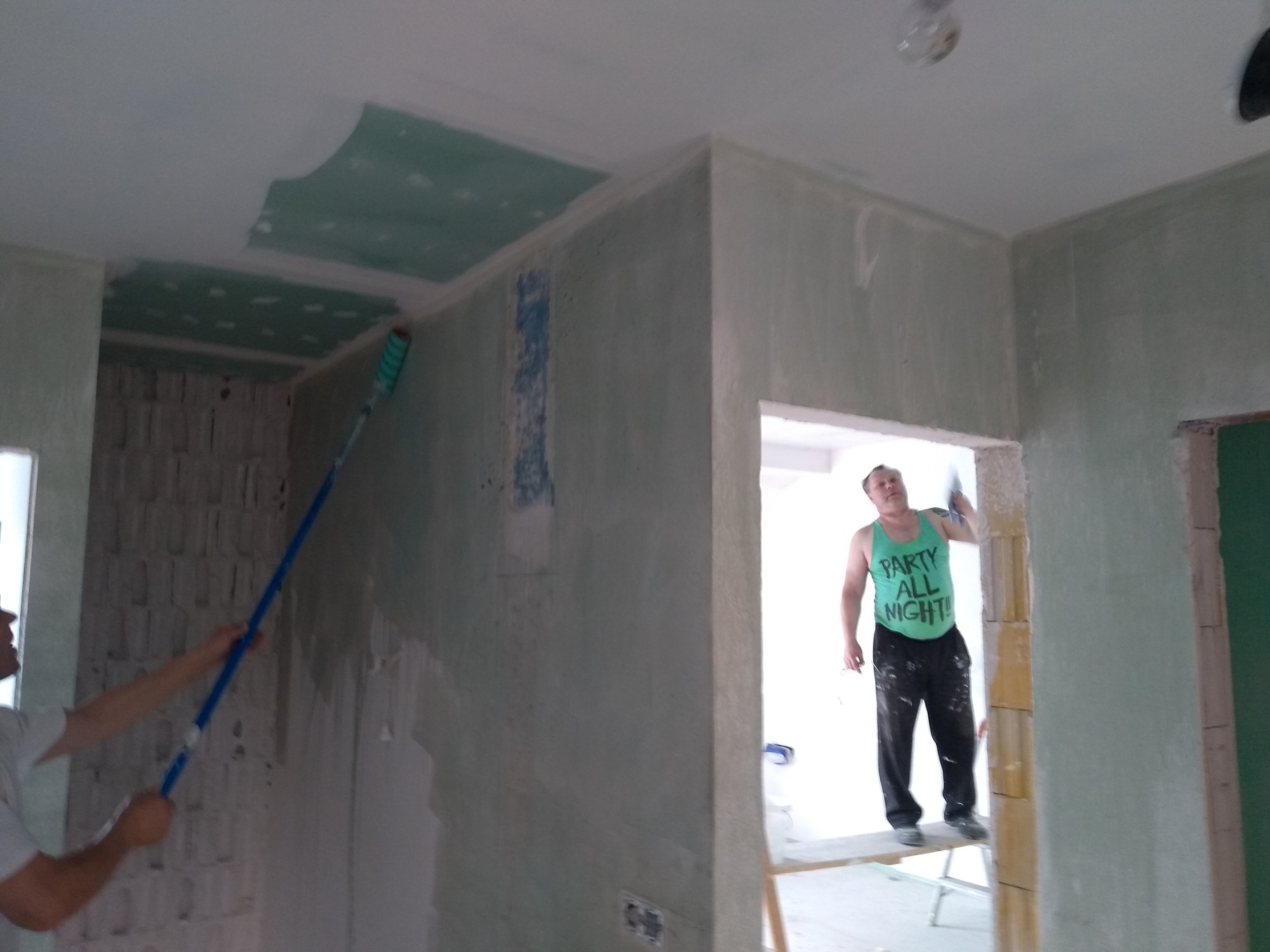 Prace wykończeniowe w pomieszczeniu: mężczyzna w zielonym podkoszulku z napisem 'PARTY ALL NIGHT' stoi na rusztowaniu i wygładza ścianę, drugi mężczyzna maluje sufit wałkiem na długim kiju...