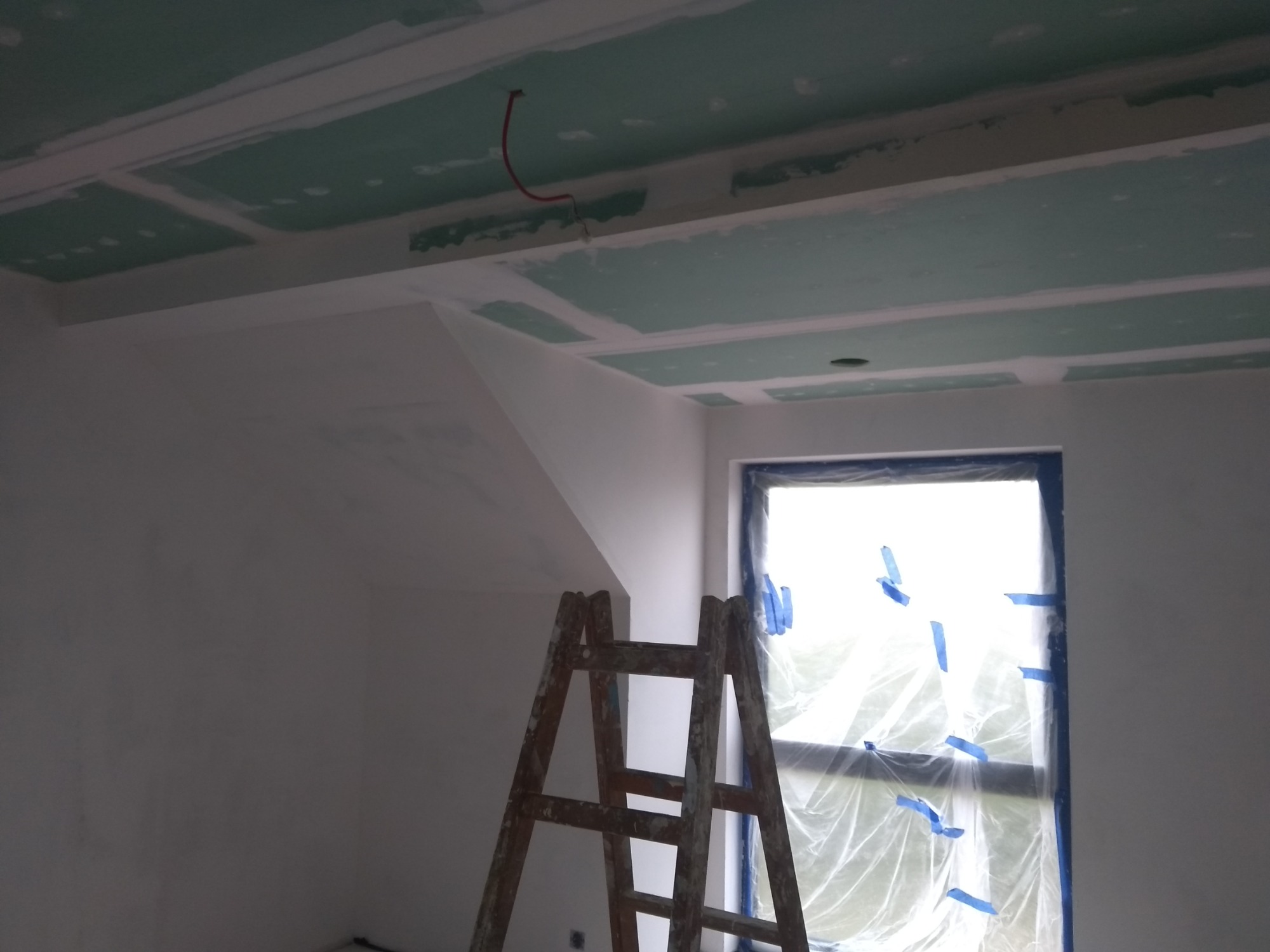 Wnętrze w trakcie prac wykończeniowych: sufit z płyt gipsowo-kartonowych, częściowo wyszpachlowany, widoczna drabina oparta o ścianę, okno zabezpieczone folią i taśmą malarską.