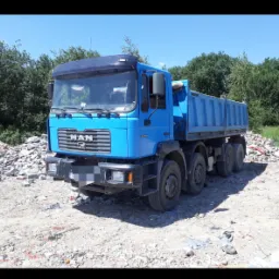 Niebieska ciężarówka MAN na placu z gruzem po rozbiórce, z zaroślami w tle.