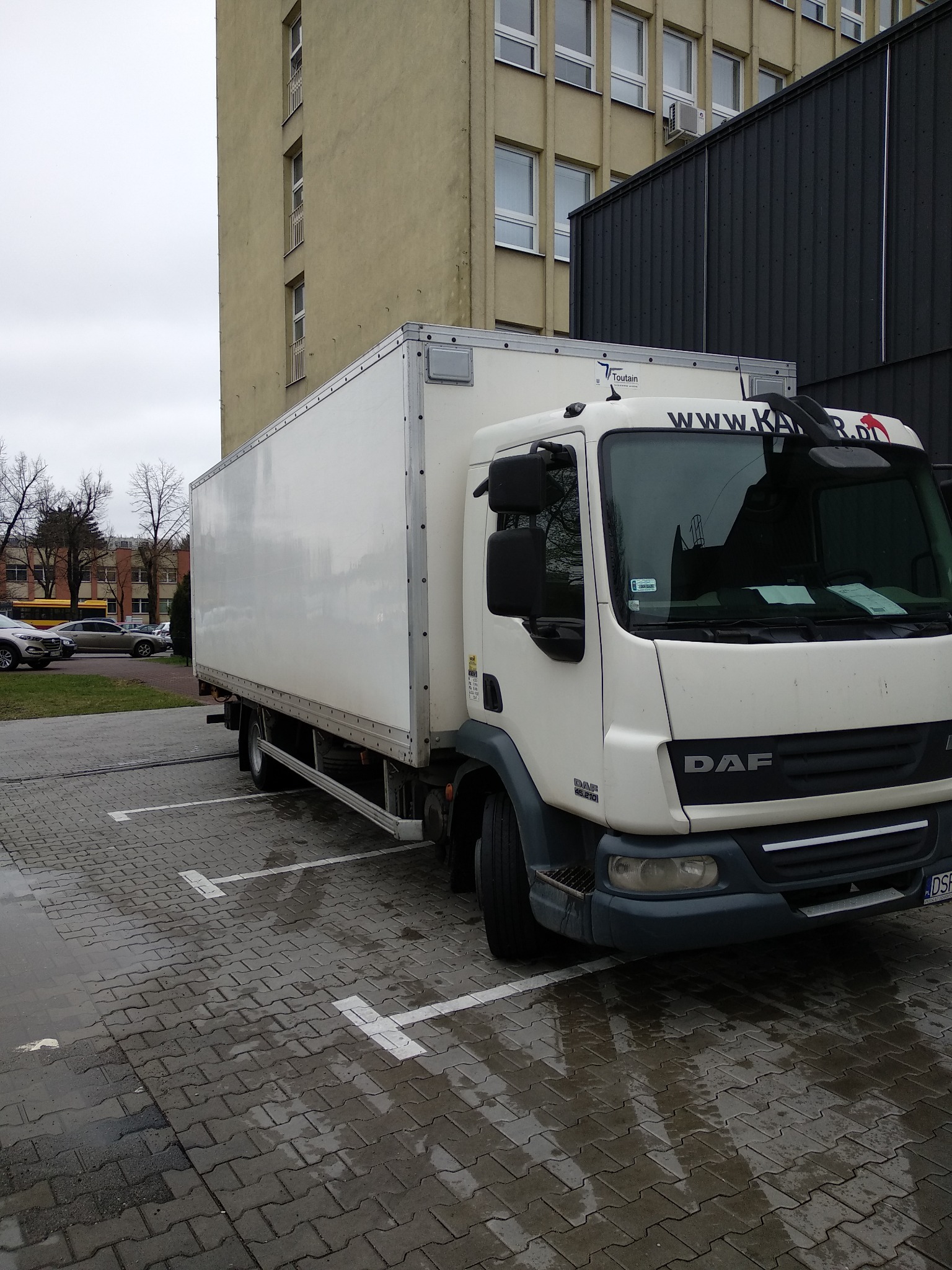 Biała ciężarówka DAF zaparkowana na mokrym parkingu z kostki brukowej, widoczny budynek w tle.