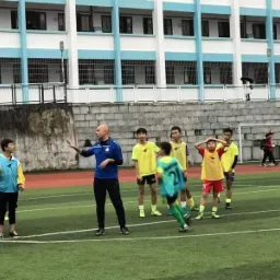 Trener piłki nożnej instruuje grupę młodych sportowców na zielonym boisku, z widocznym budynkiem szkolnym w tle.