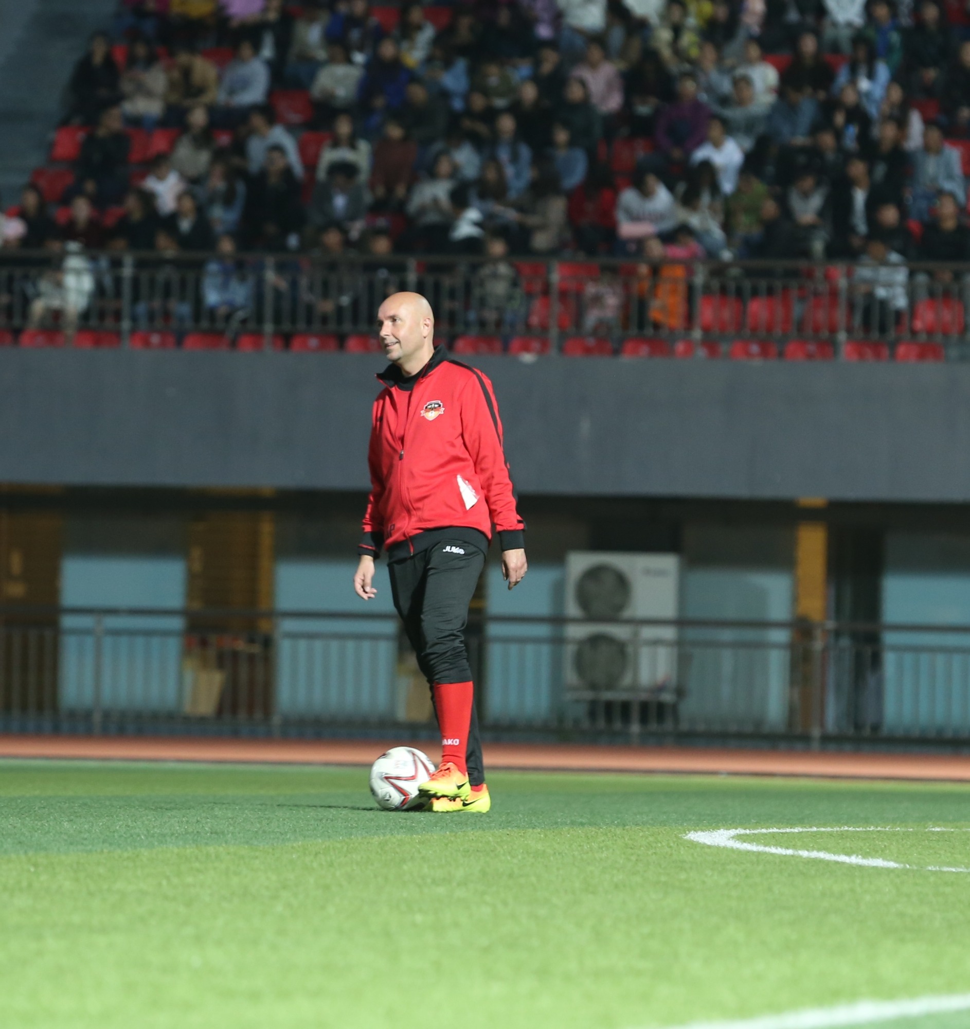 Łysy mężczyzna w czerwonej bluzie sportowej i czarnych spodniach stoi na zielonym boisku ze sztuczną trawą obok piłki nożnej, w tle trybuny z publicznością.