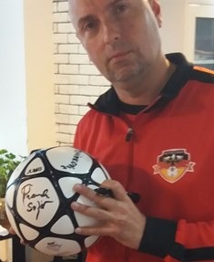 Mężczyzna w czerwonej bluzie z logo drużyny piłkarskiej trzyma piłkę nożną z autografami i pisze kolejny autograf czarnym markerem.