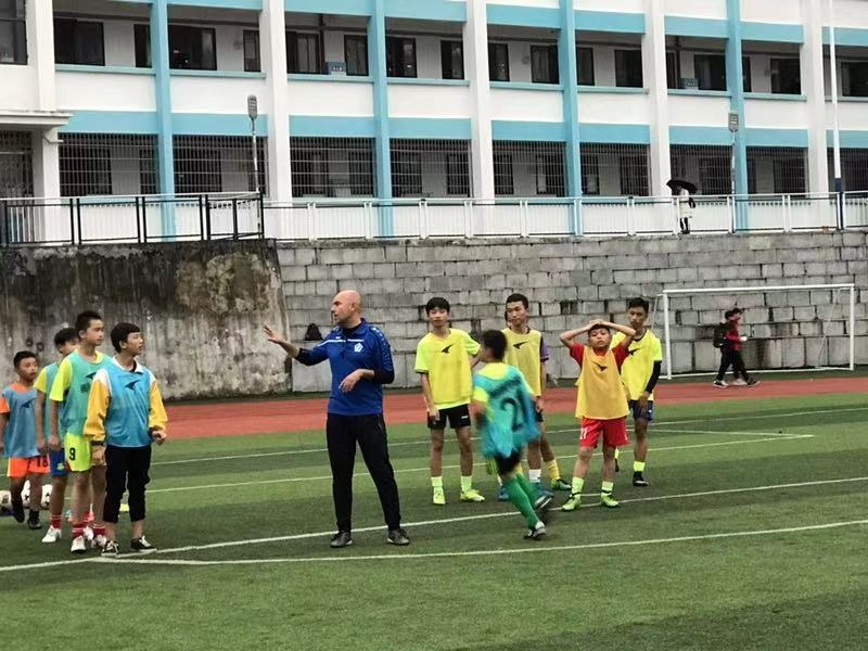 Trener piłki nożnej instruuje grupę młodych sportowców na zielonym boisku, z widocznym budynkiem szkolnym w tle.