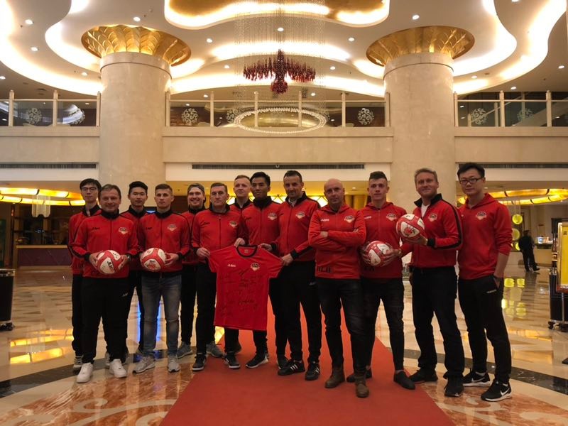 Grupa sportowców w czerwonych dresach pozuje z piłkami i koszulką z autografami w eleganckim holu hotelowym z marmurową podłogą i kolumnami.