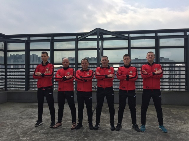 Grupa sześciu mężczyzn w czerwono-czarnych dresach z logo, stojących na tle szklanej balustrady i miejskiego krajobrazu.