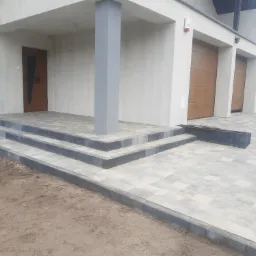 Nowoczesny podjazd i schody wejściowe do domu wyłożone jasną kostką brukową z ciemnoszarą obwódką, widoczne dwie bramy garażowe i drzwi wejściowe z oryginalnym wzorem.