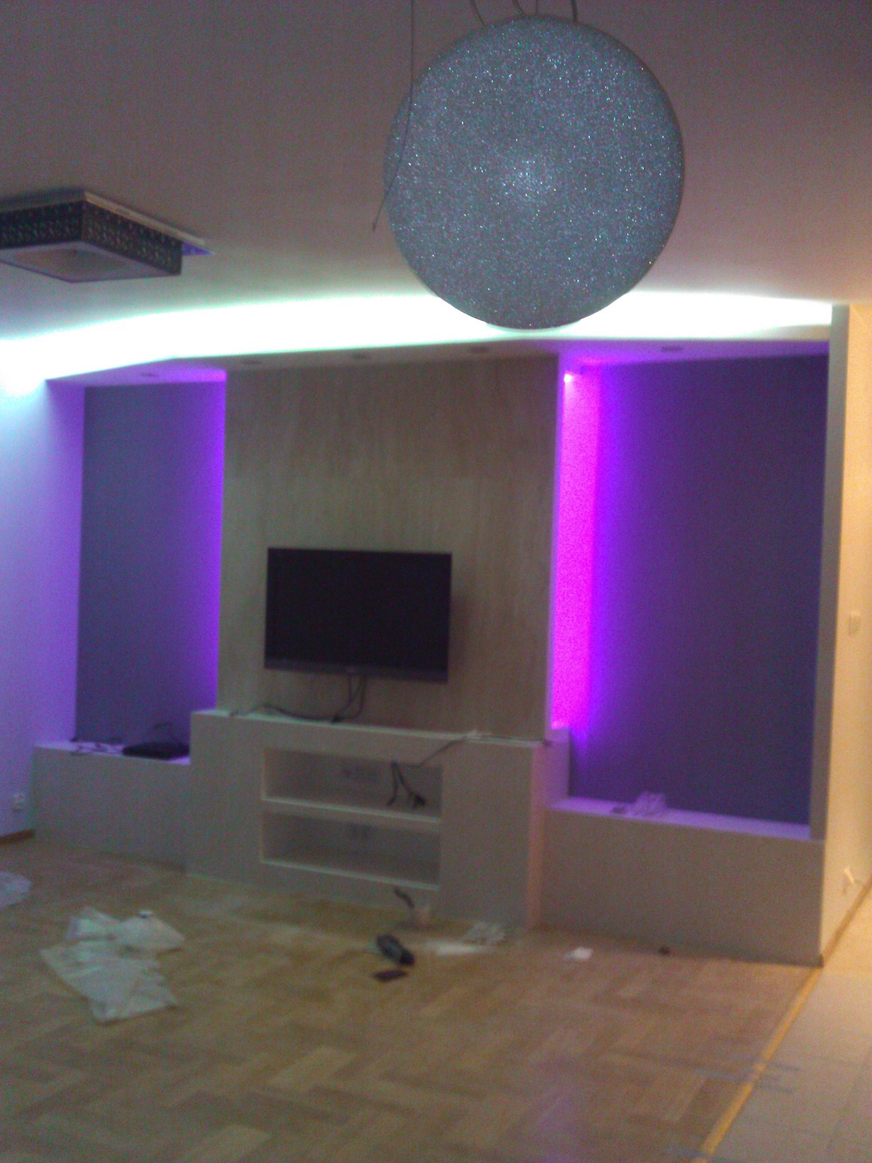 Salon z podświetlanymi na fioletowo wnękami ściennymi, zabudową pod telewizor i okrągłą lampą sufitową, widoczne ślady po remoncie na podłodze.