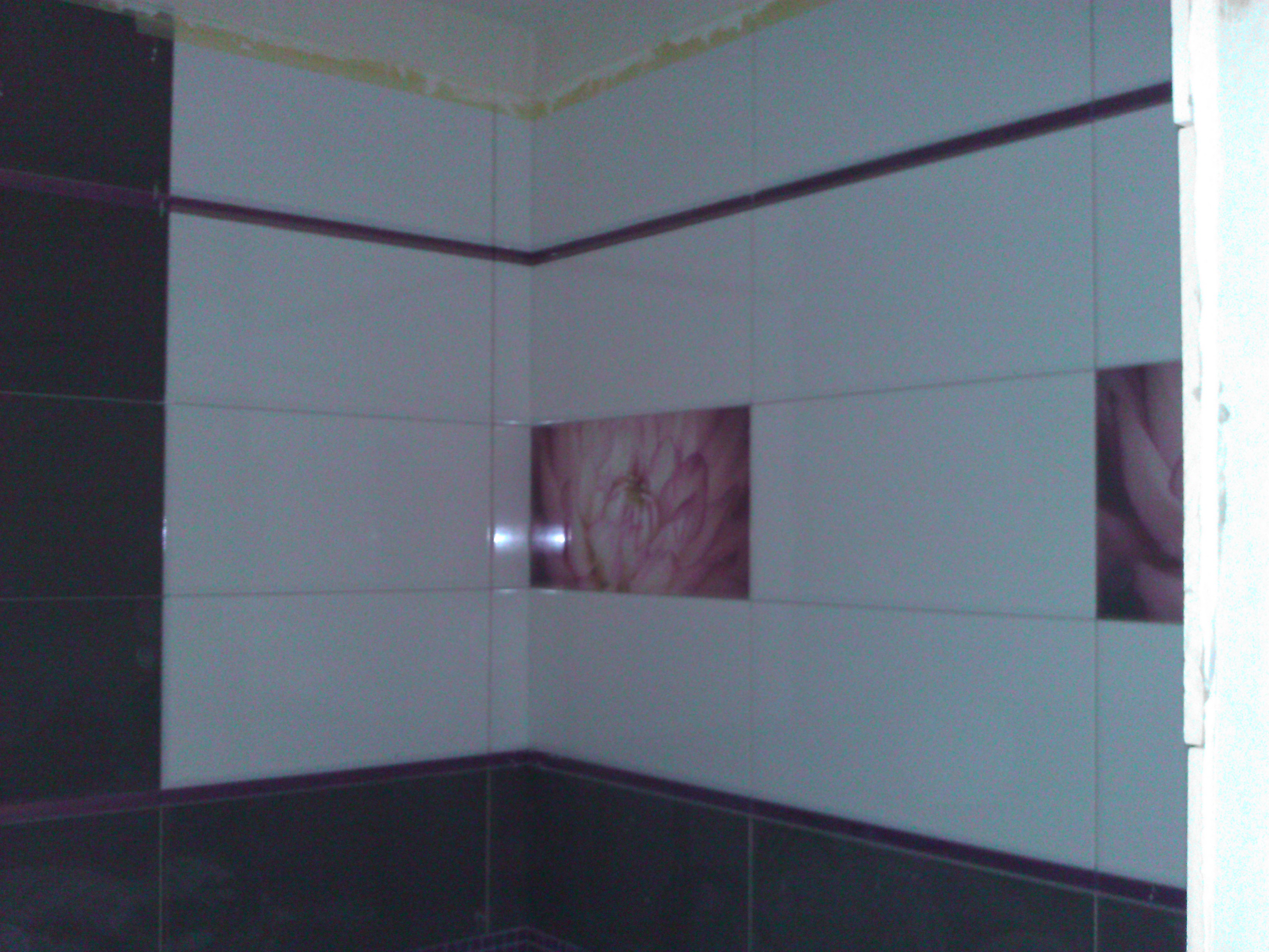 Wykończony narożnik łazienki z szarymi i ciemnografitowymi płytkami ceramicznymi, dekorowany pasem z bordową listwą i motywem kwiatowym na wysokości wzroku.